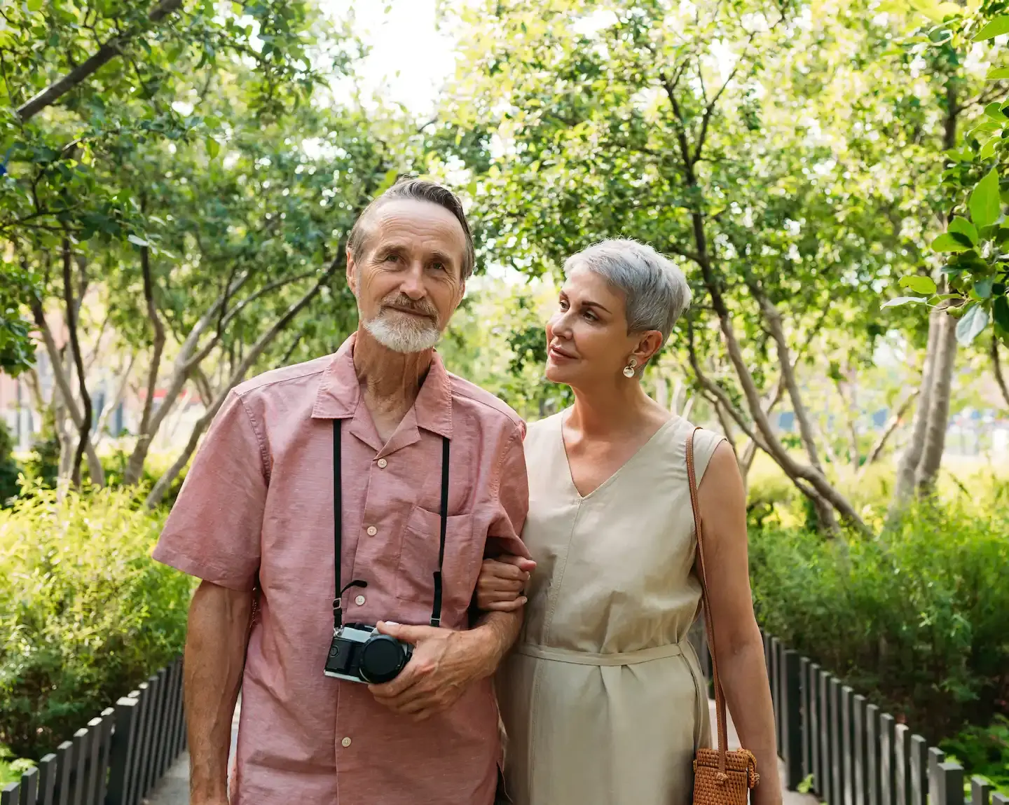 Older couple walking through a park. The man holds a camera that is strapped around his neck.